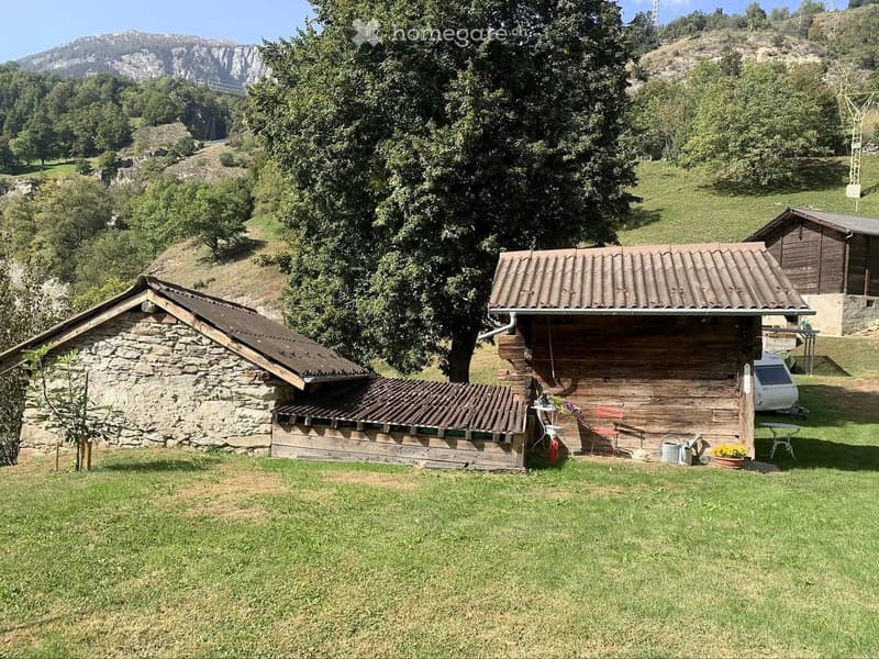 A rural outdoor area with a wooden cabin, stone structure, and a small table with chairs and a flower pot.