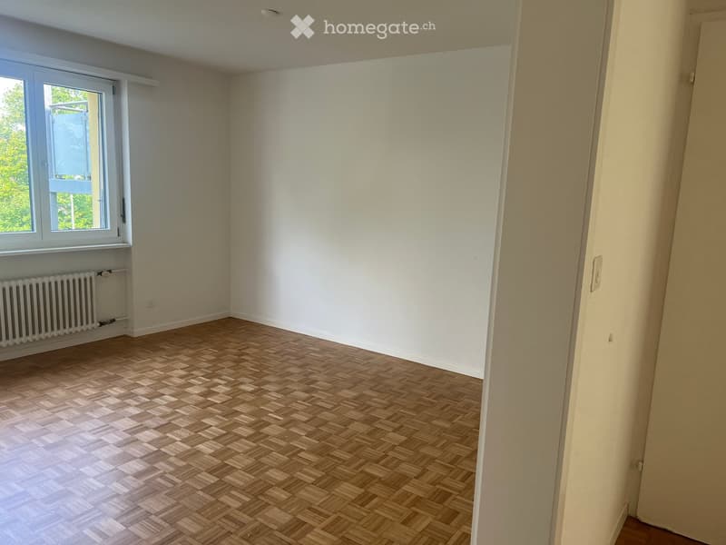 Empty room with wooden herringbone flooring, white walls, and a window with a radiator below it.