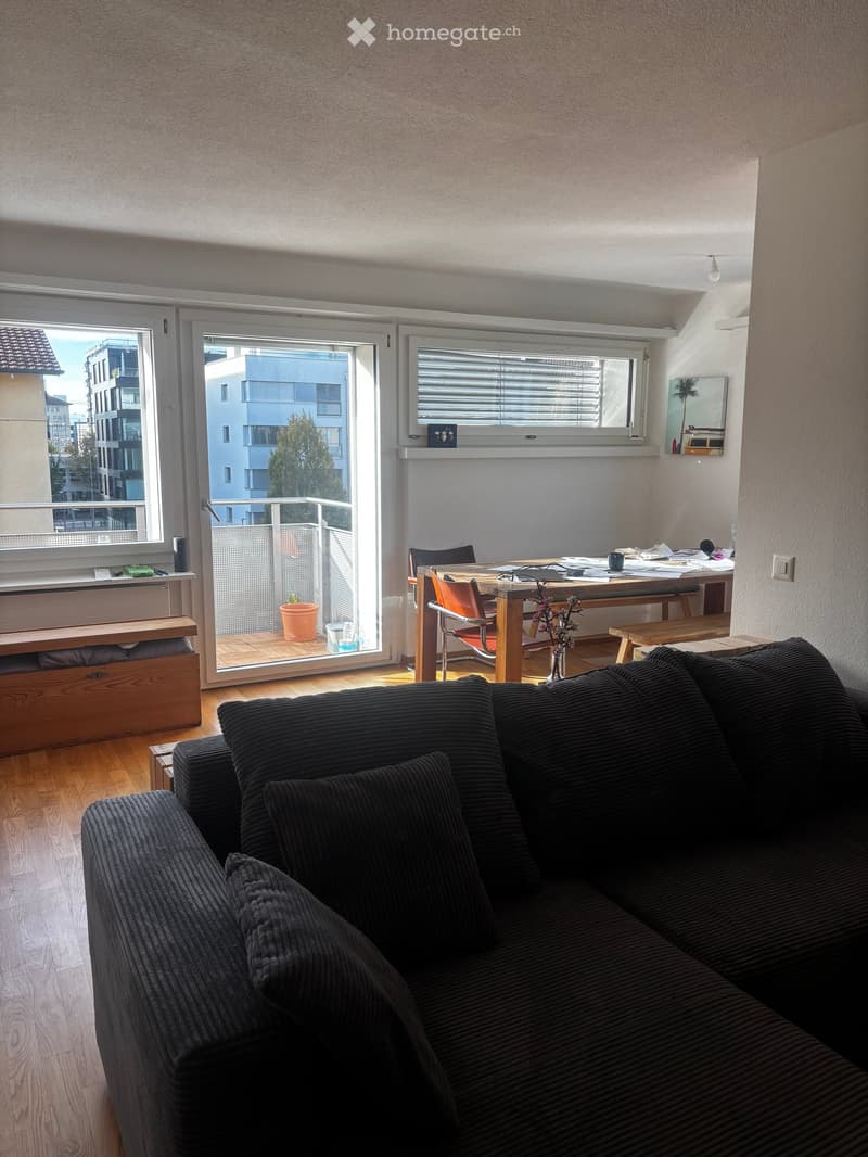 A modern living room with a dark grey sofa, a wooden dining table with chairs, a vase with flowers, and a balcony view of the city.
