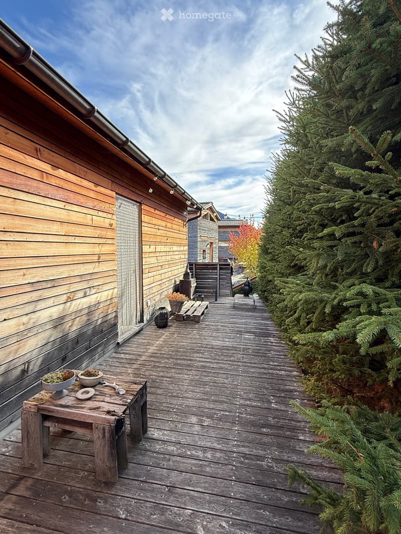 Wooden deck with small table, bowls, and potted plants, adjacent to a wooden building with a grey door and string lights.