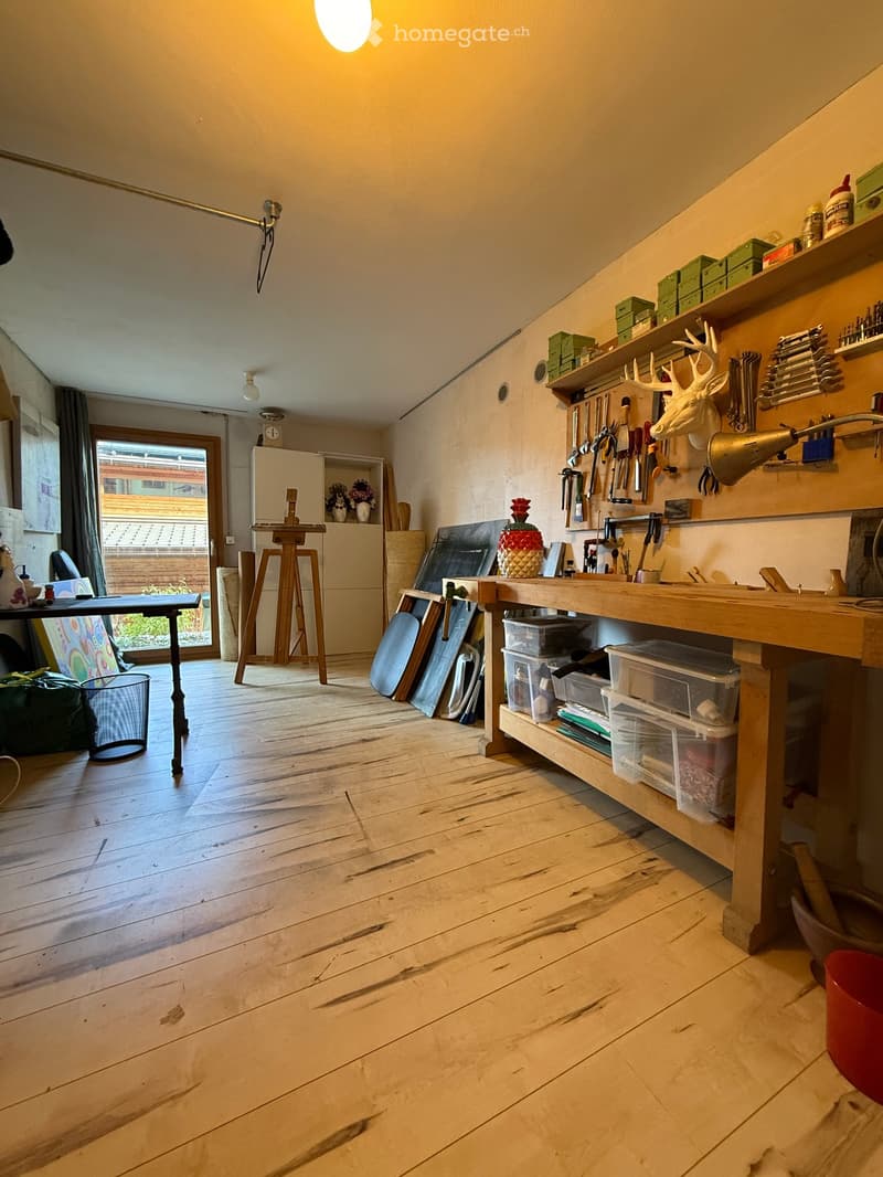Well-lit workshop with wooden floor, large workbench, shelves with tools, deer head, and various art supplies.