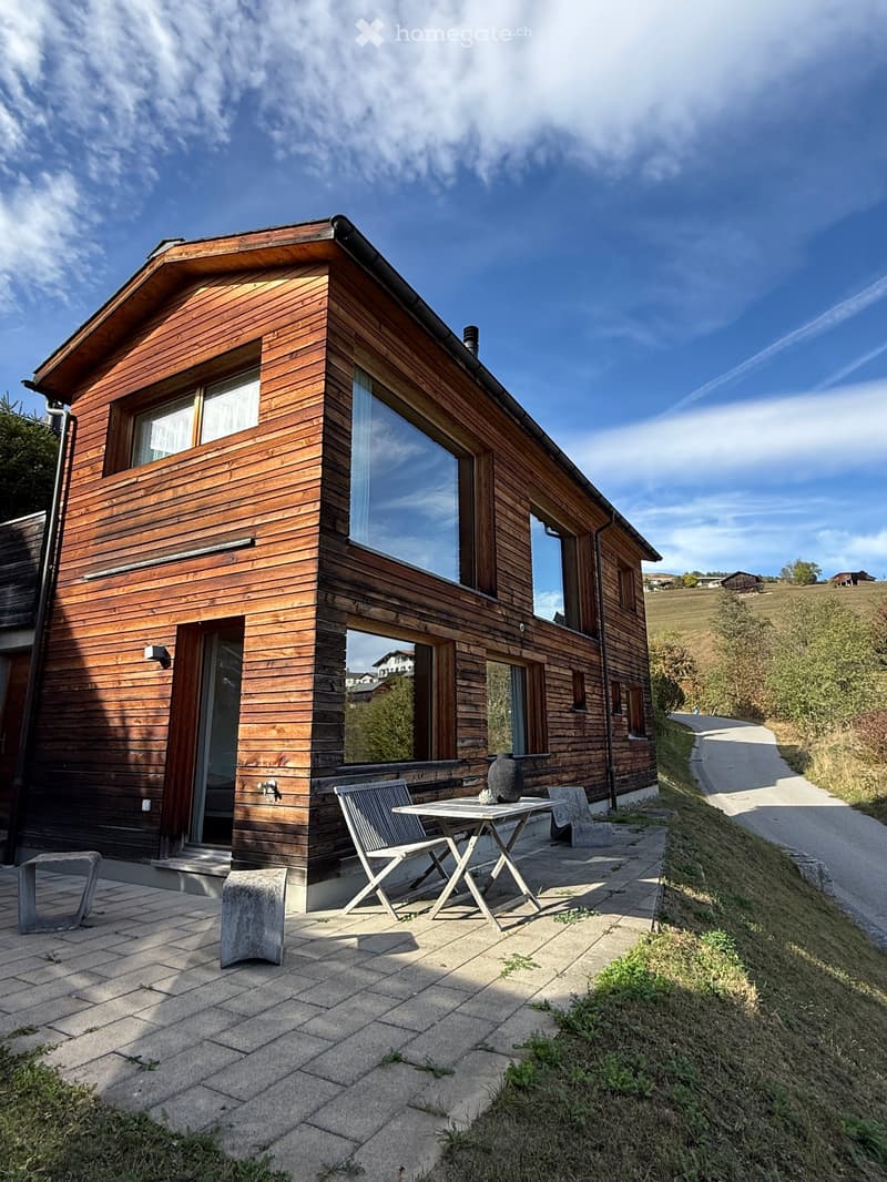Modern wooden house with large windows and a raised ground floor, featuring a patio with a table and chairs.