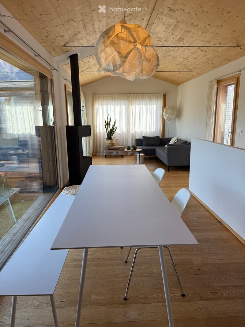 Open-plan living and dining area with wooden flooring, large windows, a modern fireplace, a dining table with chairs, a sofa, and a coffee table.