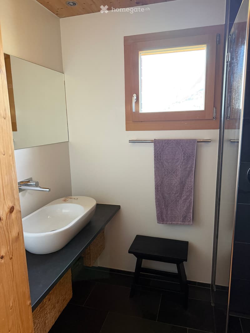 Modern bathroom with a white bathtub on a dark countertop, a small black stool, a purple towel on a rack, and a large window with wooden frame.