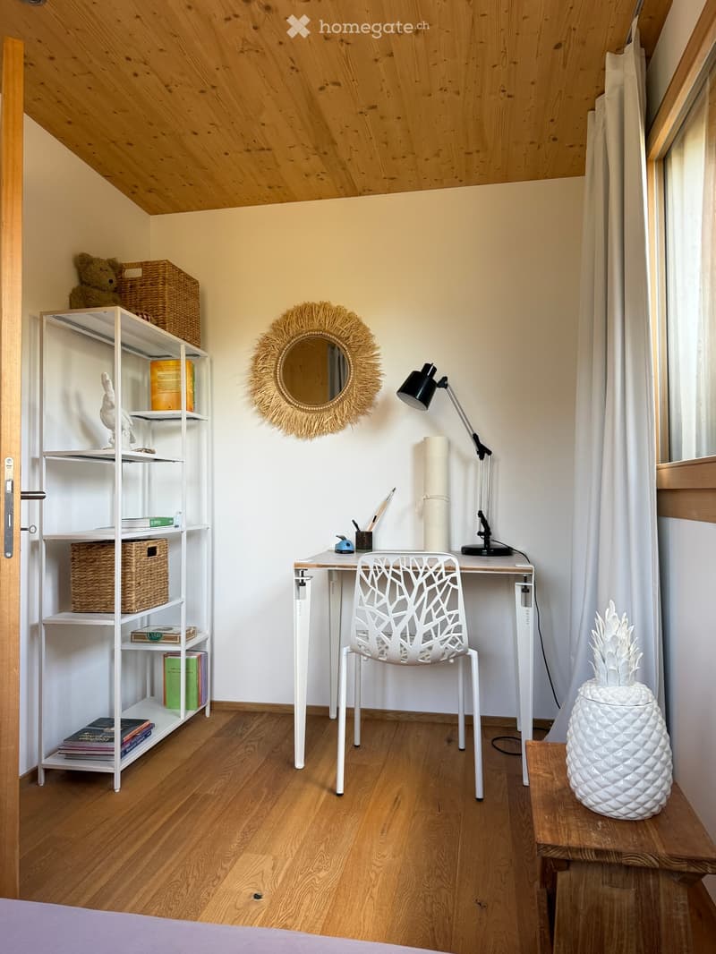 A small, well-lit room with wooden flooring, a white desk, a modern white chair, a shelf with books and baskets, a round mirror with a woven frame, and a decorative pineapple on a small table.