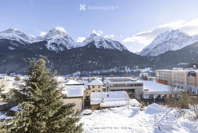 A snowy village with traditional houses and modern buildings, surrounded by snow-capped mountains under a clear blue sky.
