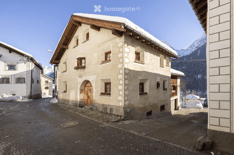 Traditional alpine building with a wooden roof, stone facade, and arched doorway, situated in a snow-covered village with mountain views.
