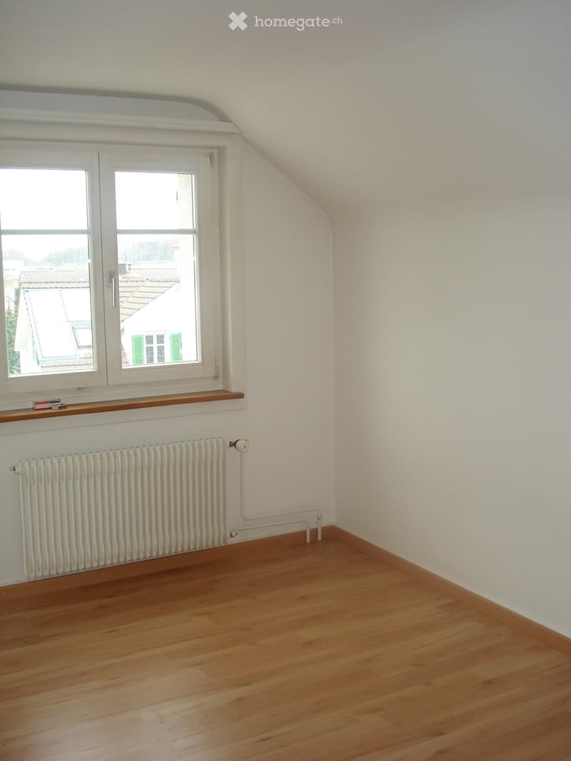 Empty attic room with wooden flooring, white walls, a radiator, and a window with a wooden shelf above it.