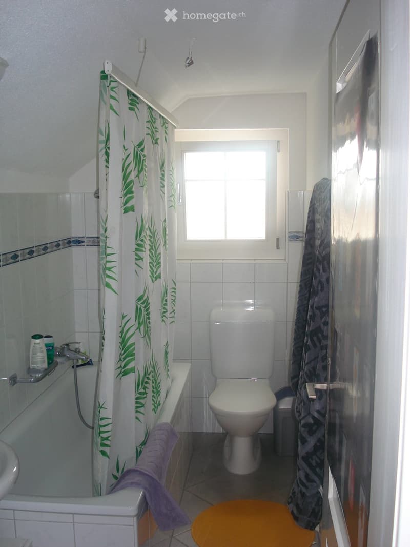 Bathroom with white tiles, a toilet, bathtub, sink, shower curtain with green leaf pattern, and a small orange stool.