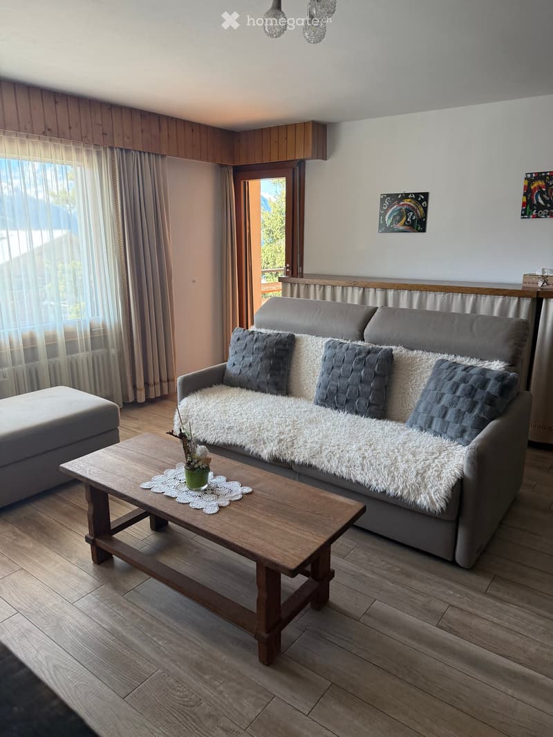 Living room with a grey sofa, white fur throw, wooden coffee table, ottoman, and a view through large windows.