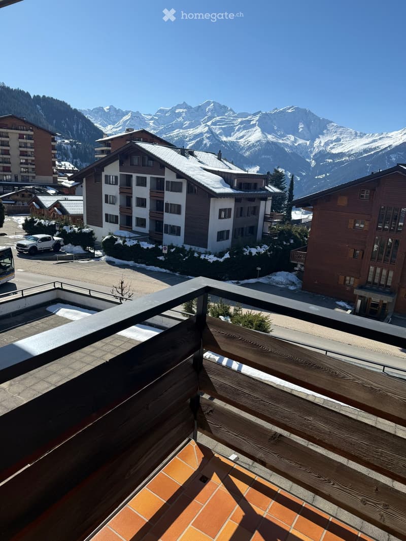 Balcony with wooden railing and orange tiled floor, overlooking a snowy town and snow-capped mountains.