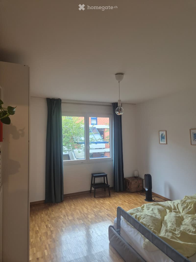 Bedroom with wooden floor, large window with curtains, bed with yellow bedding, small black stool, fan, nightstand, and picture frames on the wall.