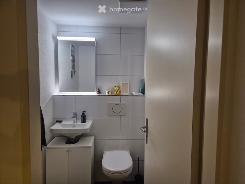 Modern bathroom with a white sink, toilet, mirror, and cabinet. The walls are tiled in light grey, and the space is well-lit with built-in lighting above the mirror.