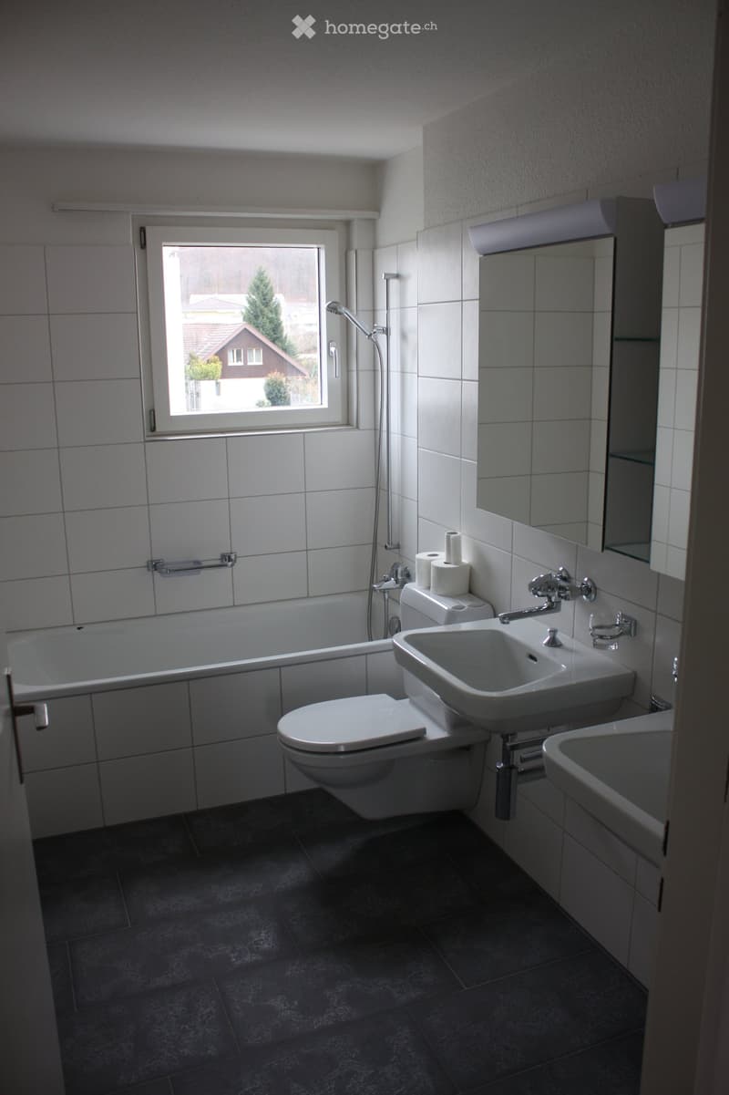 Modern bathroom with white tiles, bathtub, toilet, sink, and mirror. Features a window with a view of the outside.
