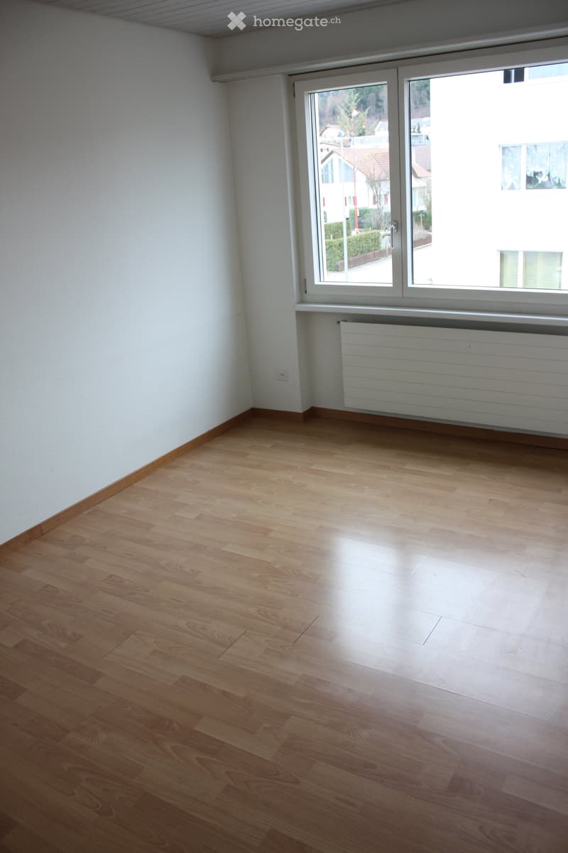 Empty room with light wooden flooring, white walls, and a large window with a radiator below it.