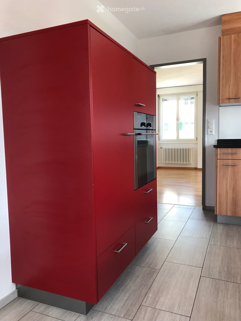 Modern kitchen with red cabinetry, stainless steel oven, and wooden flooring.