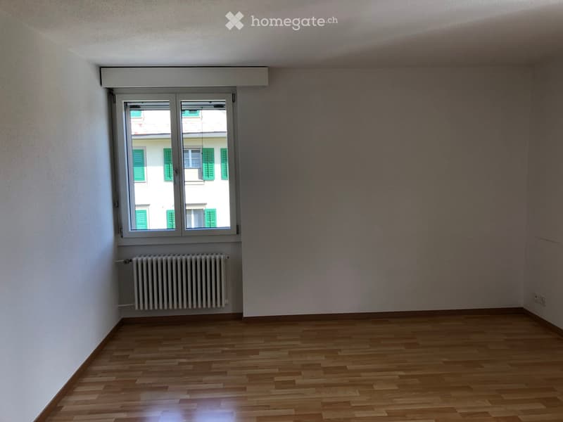 Empty room with wooden flooring, white walls, a window with green shutters, and a radiator below the window.