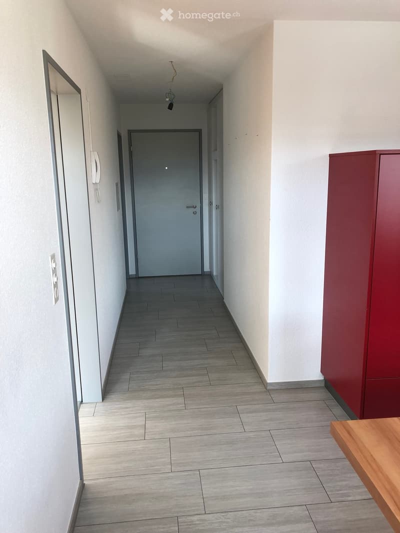 A clean, modern hallway with tiled flooring, white walls, and a wooden table on the right side. A closed door is visible at the end of the corridor.