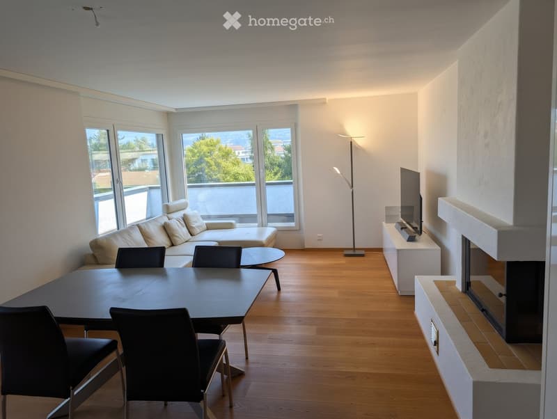 Modern living room with a white sofa, coffee table, dining table with chairs, television, floor lamp, and fireplace. Large windows offer a scenic view.