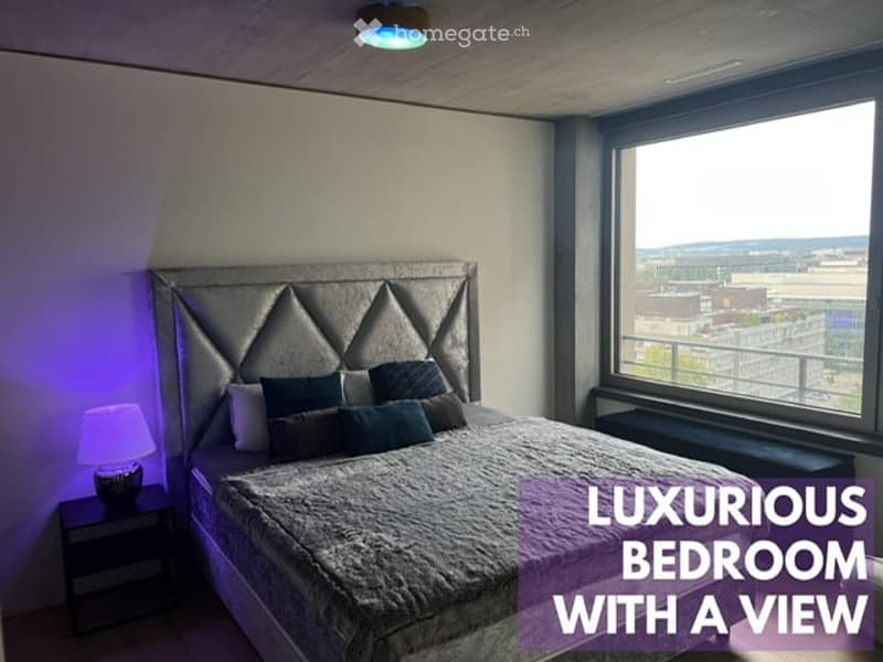 Modern bedroom with a large bed, plush headboard, nightstand with lamp, and a large window offering a city view.
