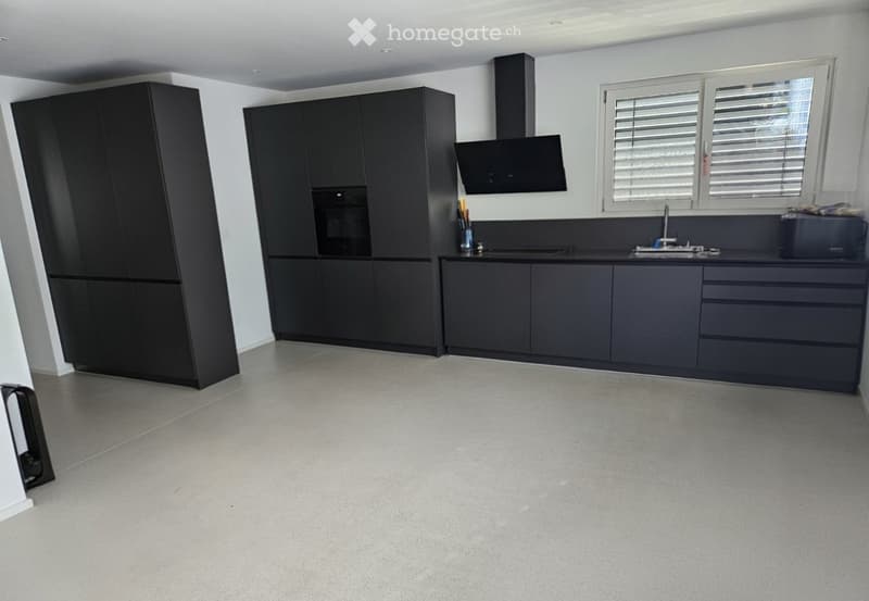 Modern kitchen with dark grey cabinets, a central island, and built-in appliances including an oven and microwave. The space features a sleek, minimalist design with a large window and a range hood above the stove area.