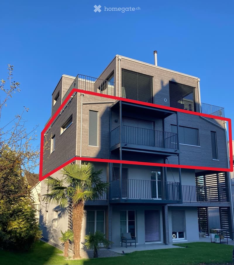 Modern two-story house with grey siding, multiple balconies, large windows, and a small garden area with a chair and table.