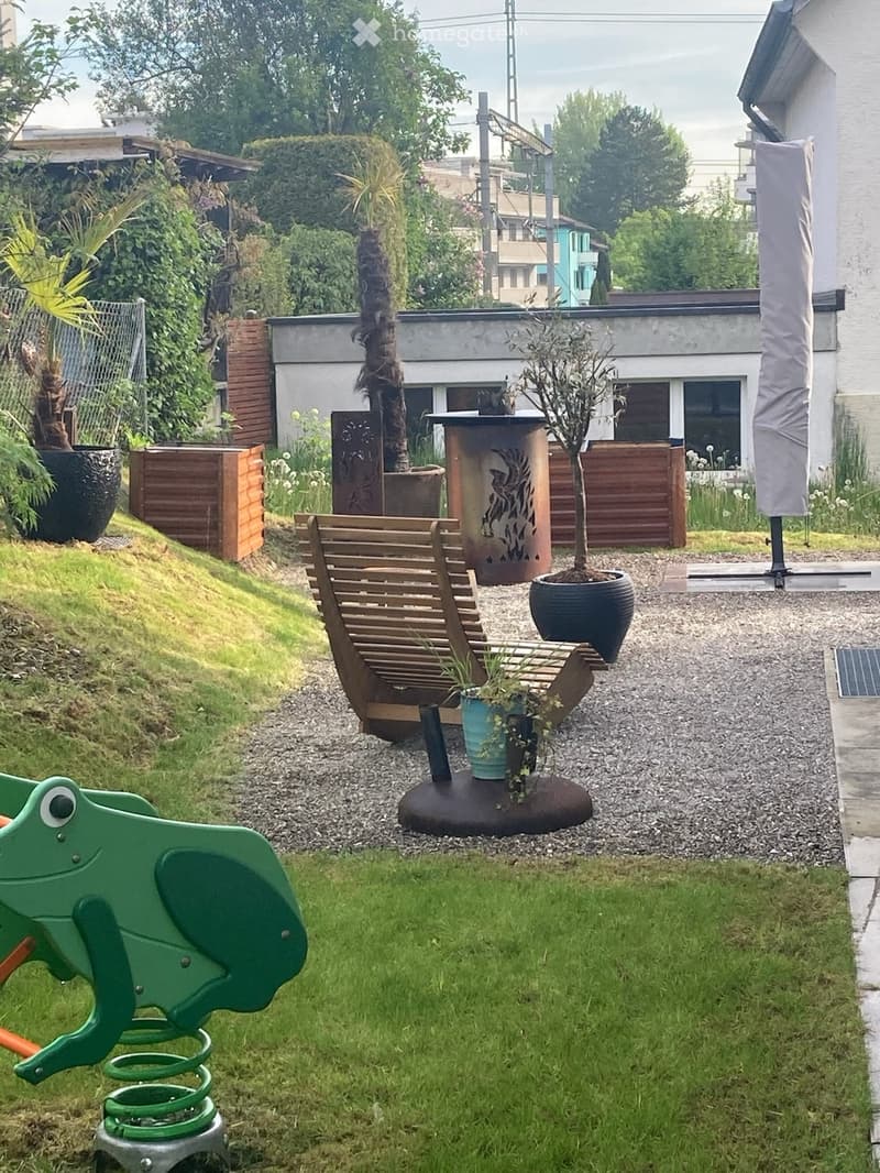 Well-maintained garden with a wooden bench, potted plants, a small grassy area, and a green frog-shaped toy on the left side.