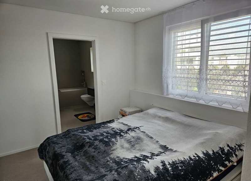 Bedroom with a large bed, patterned bedspread, white walls, and a window with white curtains. A small bathroom is visible through an open doorway, containing a toilet and a bathtub.