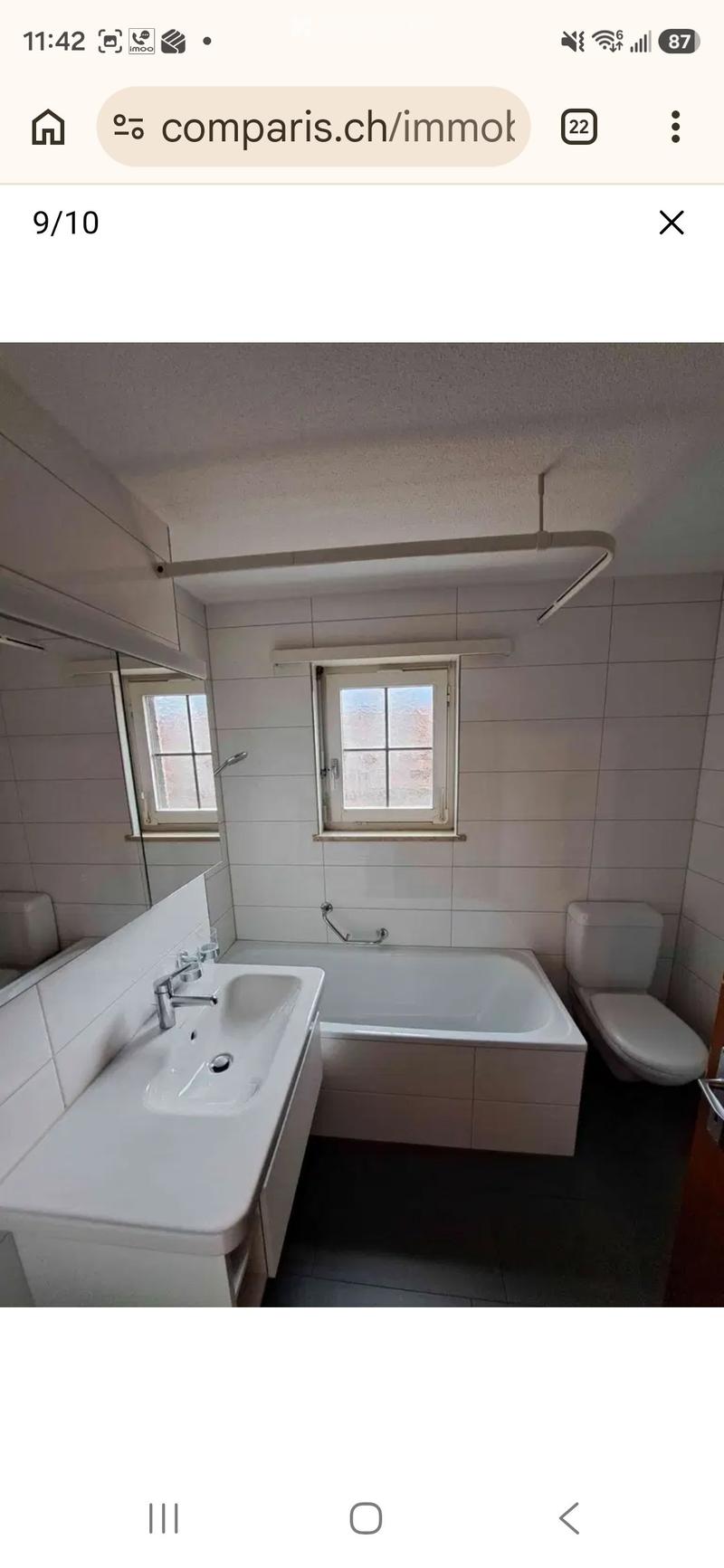 Bathroom with white tiles, a sink, bathtub, toilet, and a mirror. Two windows provide natural light.