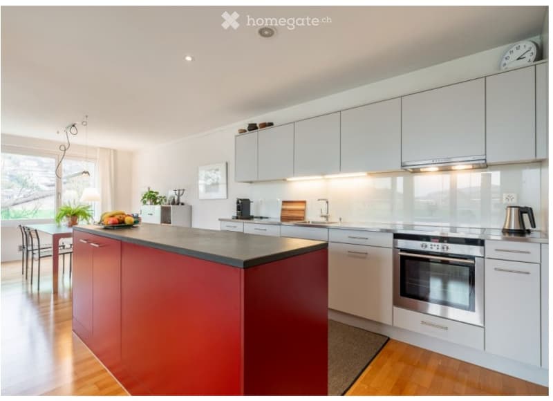 Modern kitchen with white cabinets, a red island, stainless steel appliances, and a dining area with chairs and a table.