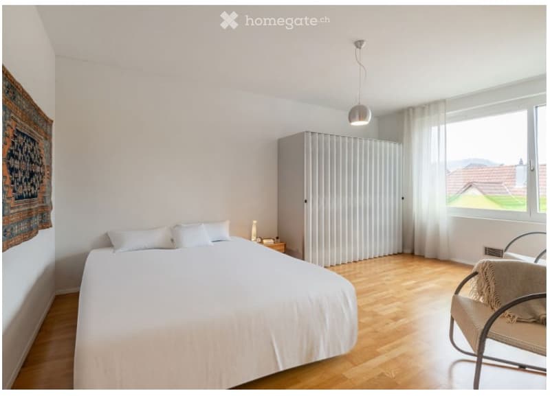 Bright bedroom with wooden flooring, a white bed with pillows, a nightstand, a chair with a throw, a wardrobe, and a decorative rug on the wall.