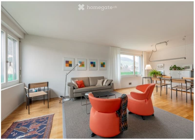 Bright living room with large windows offering a scenic view, featuring a grey sofa with colorful cushions, a coffee table, orange chairs, a floor lamp, and a dining area with a table and chairs.