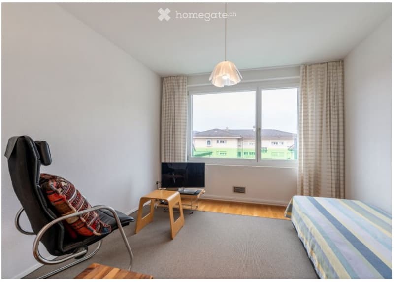 A furnished room with a bed, chair, stool, table, and television. The room has large windows with curtains and a hanging light fixture.