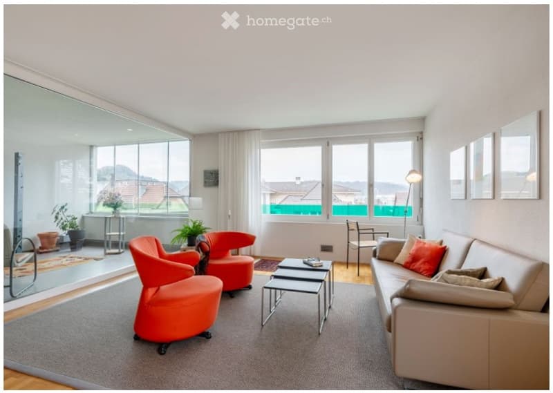 Modern living room with large windows offering a view, featuring a beige sofa with red pillows, red armchairs, a coffee table, floor lamp, and decorative plants.