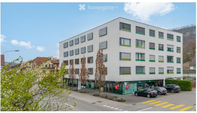 Modern multi-story building with white facade and green window frames, located in an urban area with parked cars and a pedestrian crossing nearby.