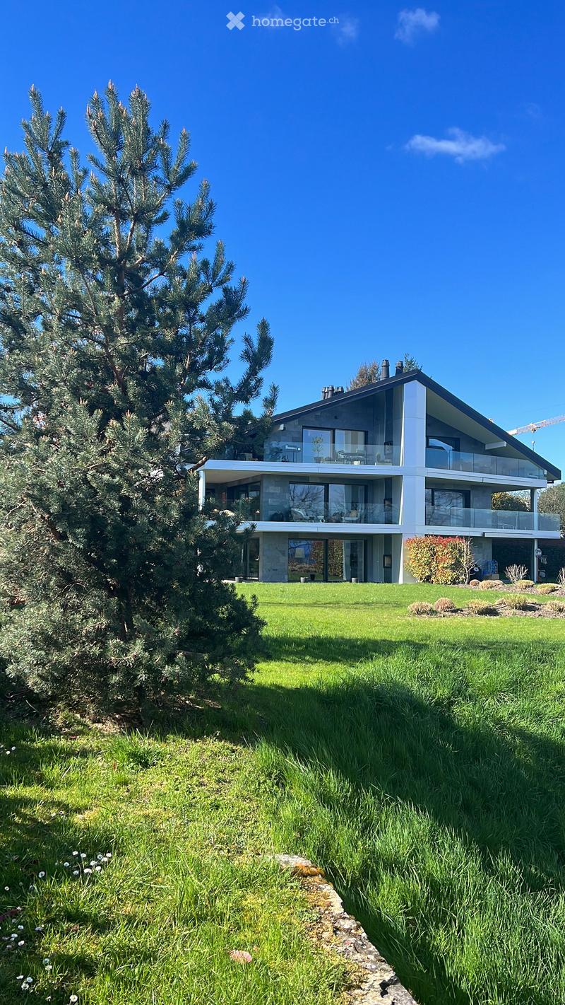 Modern two-story house with large windows, a raised ground floor, and a well-maintained lawn.