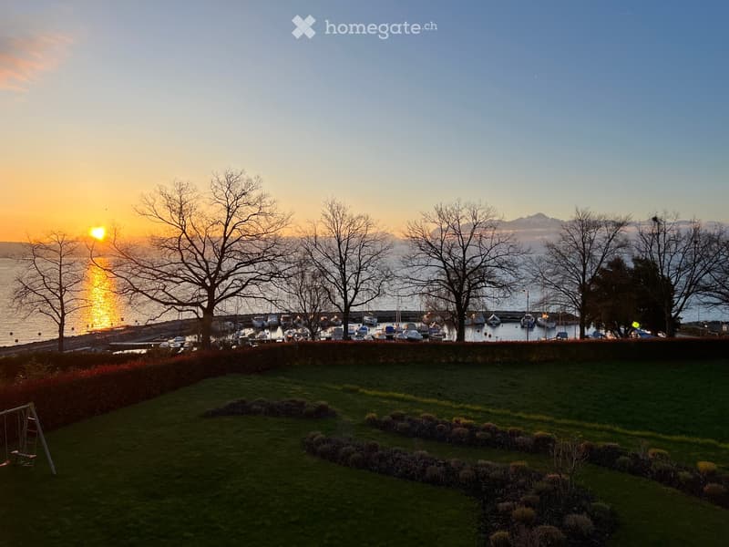 A grassy garden area with a swing set, overlooking a harbor with boats and a sunset view over the water.