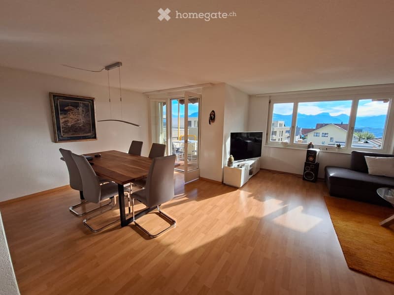Bright living and dining area with wooden flooring, large windows, a sofa, dining table with chairs, TV, speakers, and a pendant light.