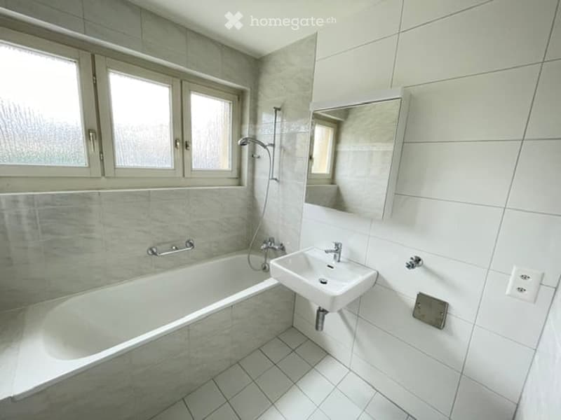 Modern bathroom with a bathtub, sink, mirror, and showerhead. The room features tiled walls and floor, with three windows providing natural light.