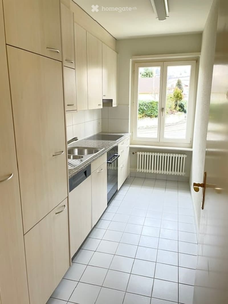 Modern kitchen with light wooden cabinets, white tiled floor, stainless steel sink, and oven. Large window provides natural light.