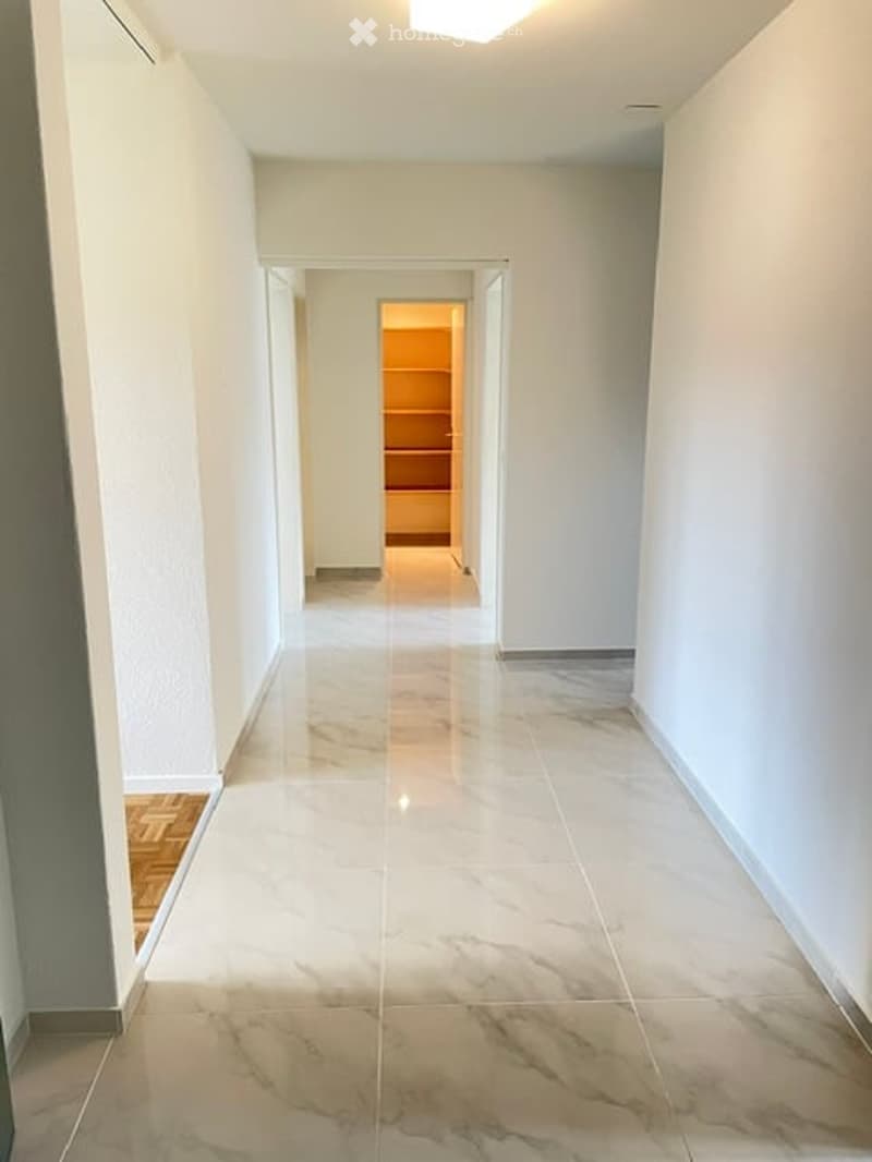 Clean, well-lit hallway with white walls and marble flooring, leading to an open doorway.