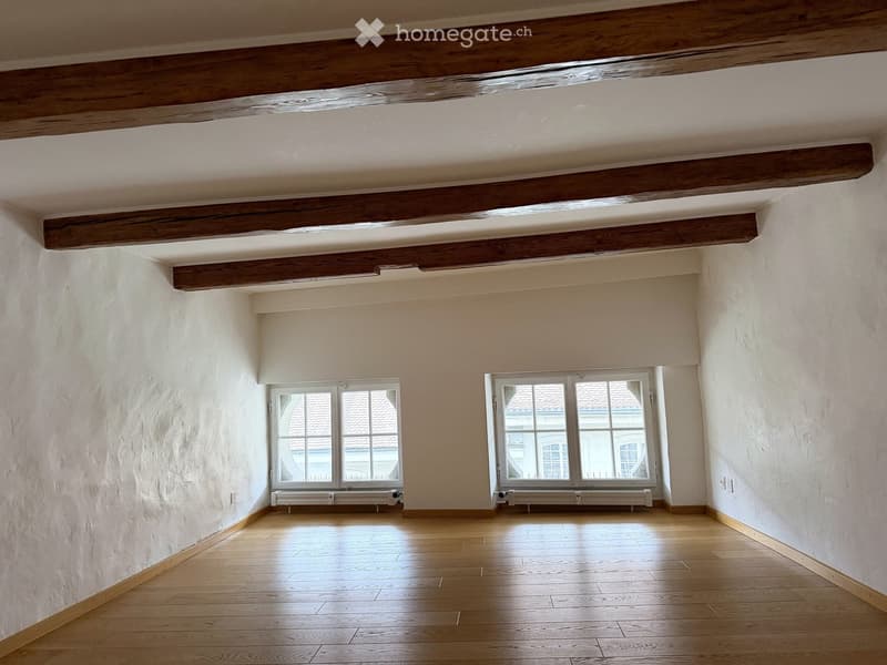 Empty room with wooden floor, white walls, exposed wooden beams on the ceiling, and two windows with white frames and radiators below them.