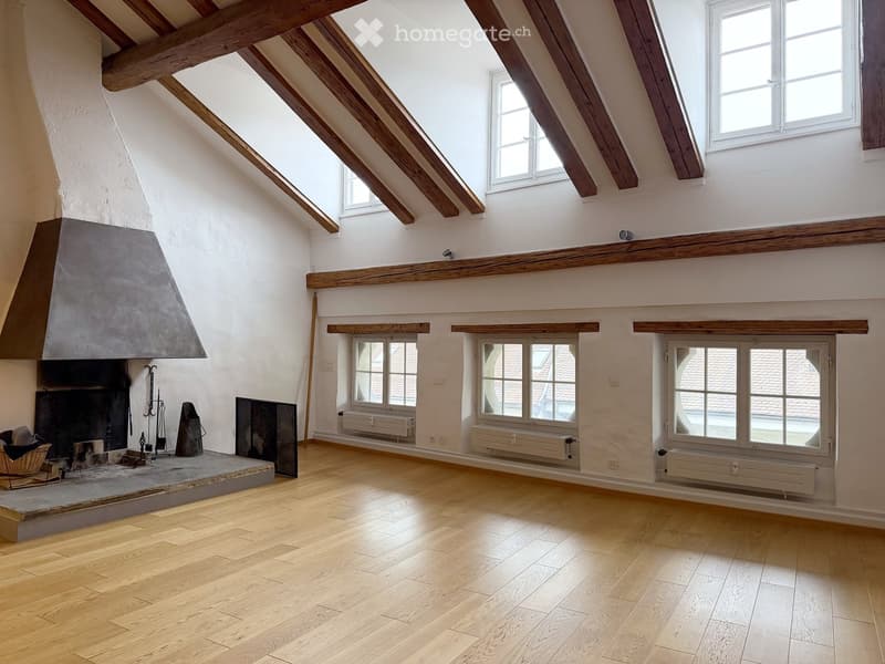 Empty attic room with wooden beams, exposed wooden floor, fireplace, and multiple windows.