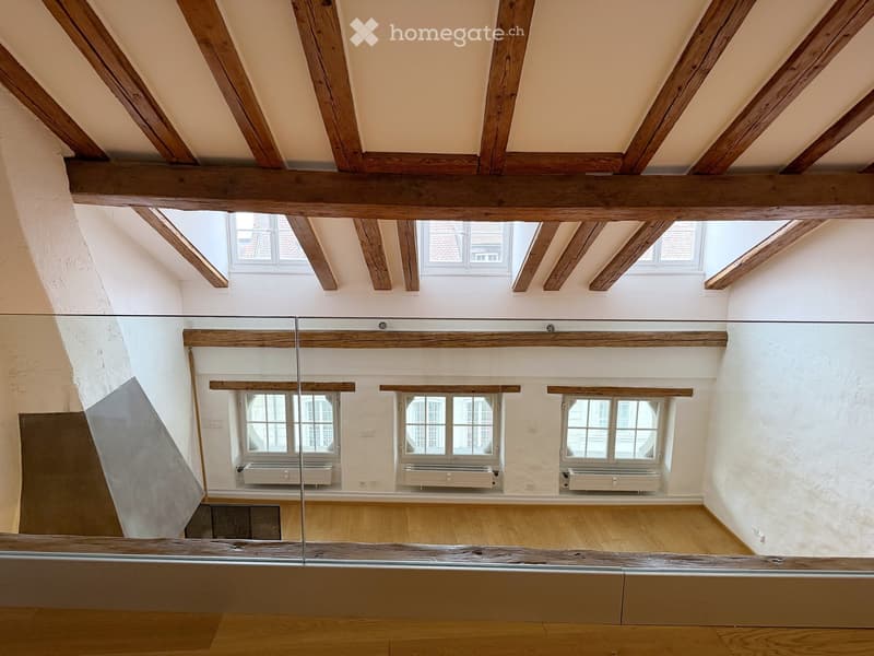 An empty room with exposed wooden beams, white walls, wooden flooring, and multiple windows, including skylights, with radiators beneath the windows.