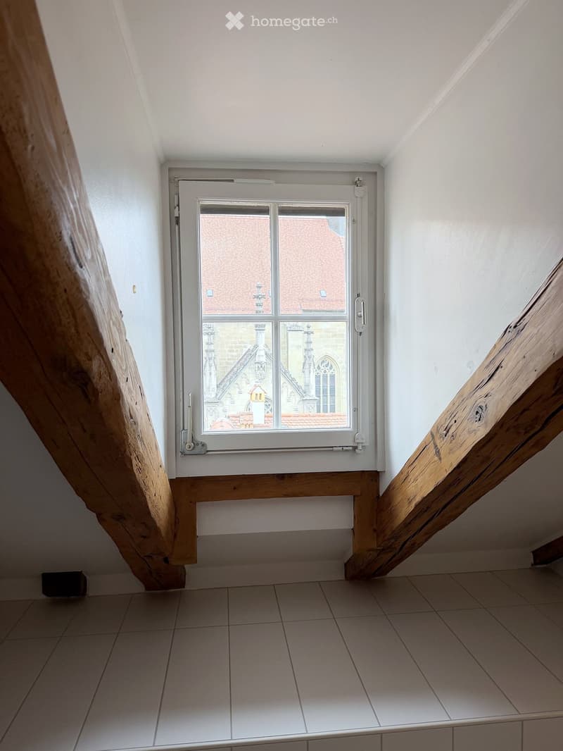 Empty room with tiled floor, white walls, and a window showing a view of a building with a red roof and architectural details.