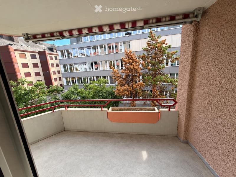 Balcony with concrete floor, red metal railing, and a planter box, overlooking a cityscape with buildings and trees.