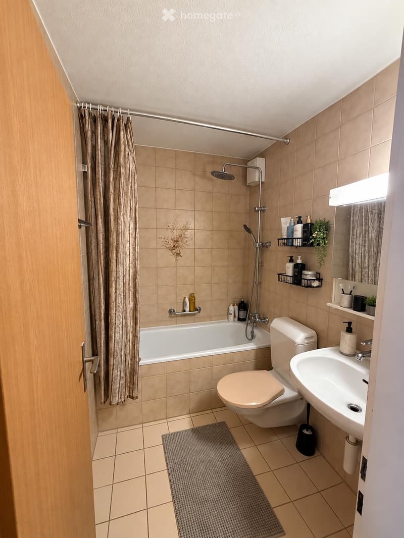 Modern bathroom with beige tiles, bathtub, toilet, sink, shower, and shelves holding toiletries and plants.