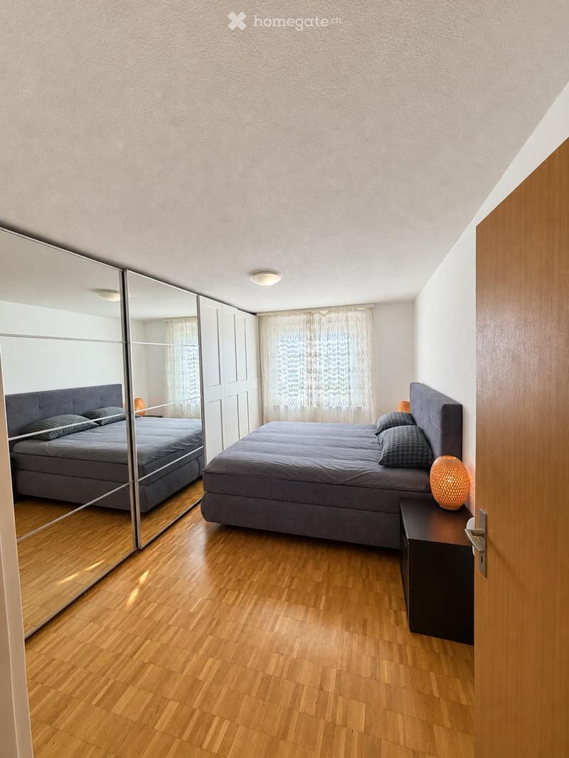 Modern bedroom with a large bed, nightstand, lamp, and a large mirror reflecting the room.