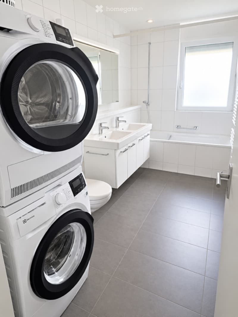 Modern bathroom with white tiles, a double sink, a bathtub, a mirror, and a stacked washer/dryer unit.