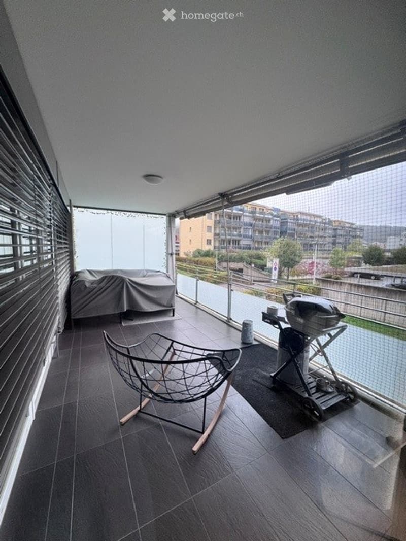 Modern balcony with dark tiled flooring, metal-framed chair, covered furniture, and a barbecue grill, overlooking a cityscape with buildings and a body of water.